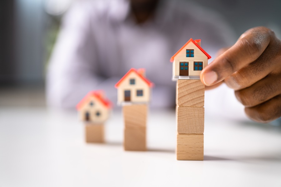 Ręka układa modele domów na drewnianych blokach, pokazując gradację cen i ryzyko rynku deweloperskiego w Bułgarii.